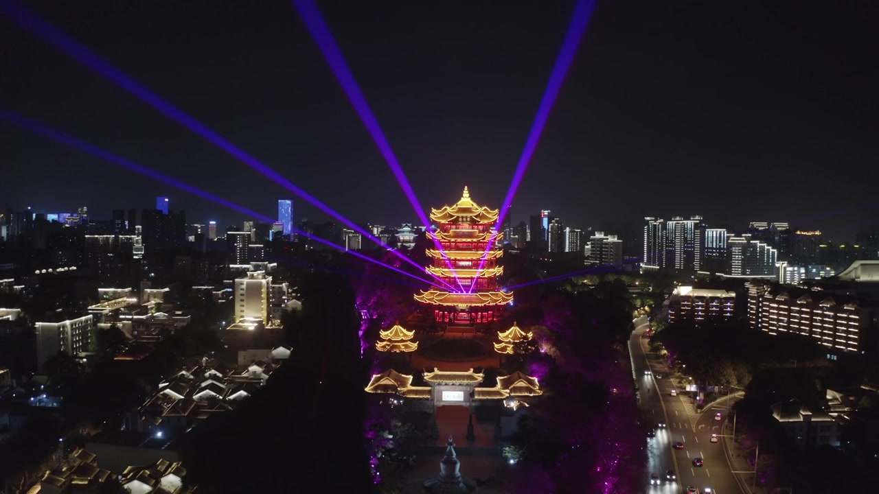Vista aérea noturna panorâmica da iluminada Torre do Grou Amarelo disparando feixes de laser ciano pelo horizonte da cidade em Wuhan, China, emoldurada por avenidas iluminadas em azul, edifícios brilhantes e tráfego rodoviário.
