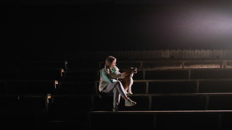 tamer seated on podium at night rubbing ears of pet playfully as bright spotlight illuminates surroundings creating calm bonding moment between human and pet under empty dark stadium setting