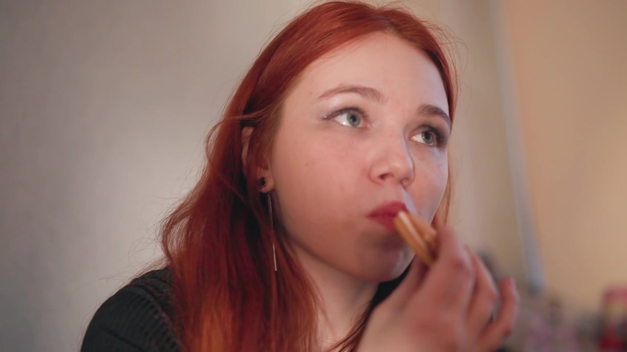 Nærbilde av en rødhåret kvinne på soverommet, som leker spådom mens hun spiser snacks. Myk bokeh-bakgrunn, varmt omgivelseslys, ærlige og ettertenksomme uttrykk, med en leppestiftberøring og fingertuppgest.