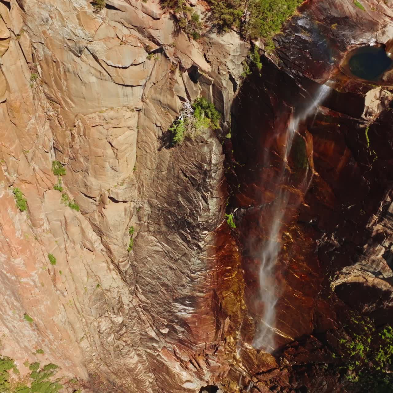 Rainbow appearing in the stream falling from huge cliff. Drone footage raising along the steep rocks of Yosemite National Park, California, USA