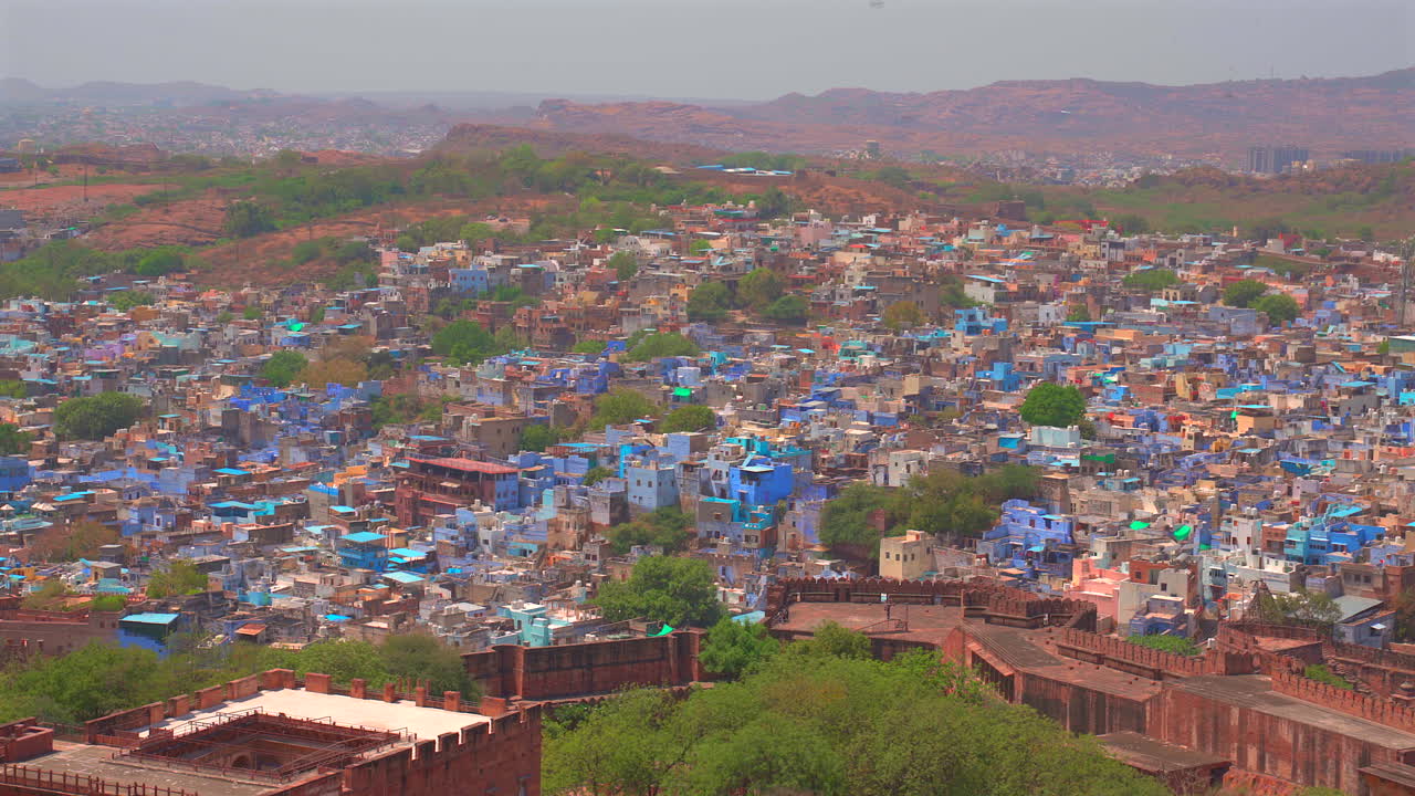 インド、ジョードプルの密集した青く塗られた家々を見下ろす広角の固定ショット。背景にはラジャスタン砂漠の険しい丘と要塞の壁が見えます。