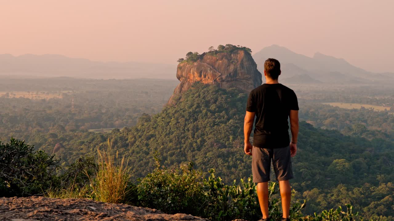 A man gazes thoughtfully at the iconic Sigiriya Rock as the sun rises over the lush landscape of Sri Lanka.
