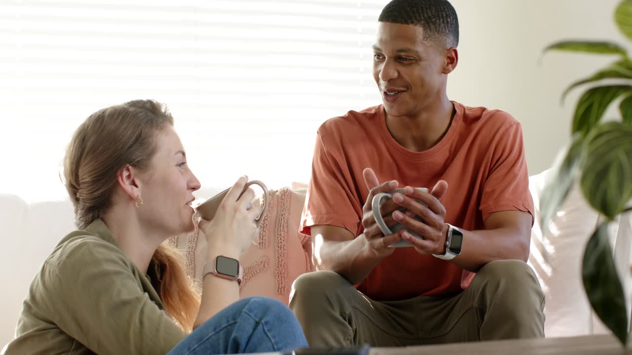 Diverse friends chatting on couch in bright living room, man speaking, woman lifting mug, sipping