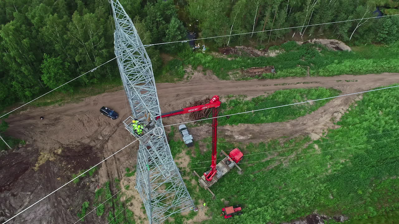 Aerial top down shot of industrial worker lifting by crane repairing electricity pylon in woodland