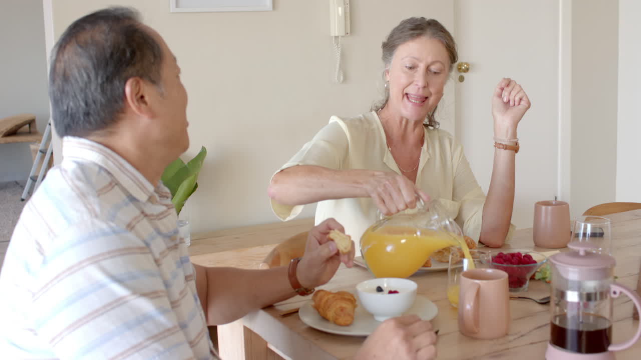 Pouring orange juice, senior woman enjoying breakfast with senior man at table
