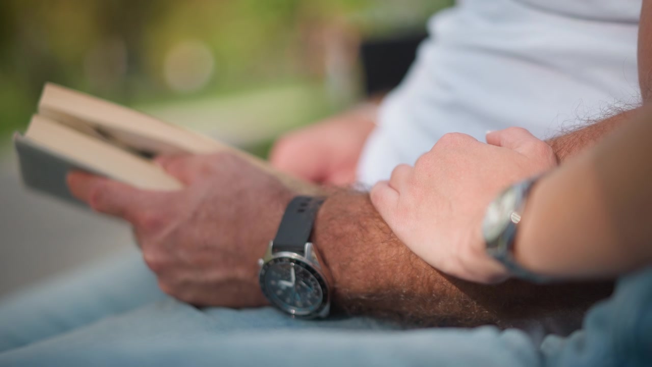 Close up of two people adult man and young girl with watches on their wrists holding hands while older person reads from books in soft natural light. intimate gesture suggests connection