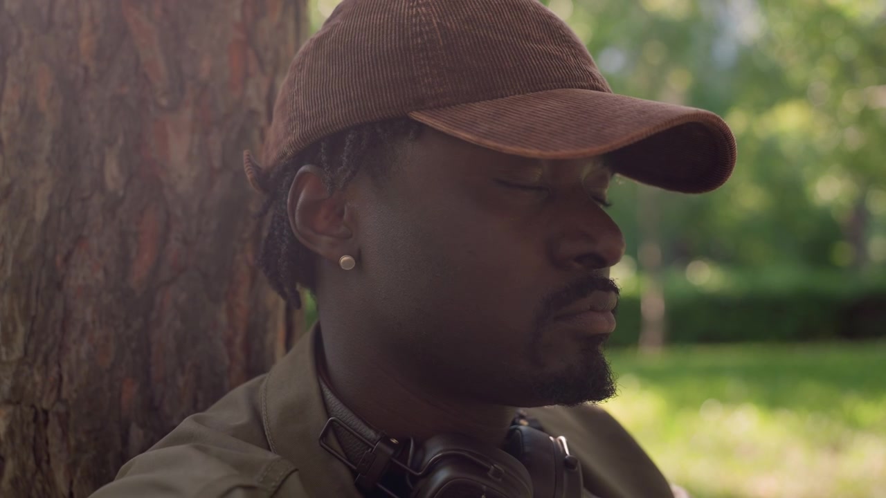 Close up of man adjusting brown cap while wearing headphones around neck standing near tree in park setting with green foliage and soft daylight. Man has earring visible on ear and jacket