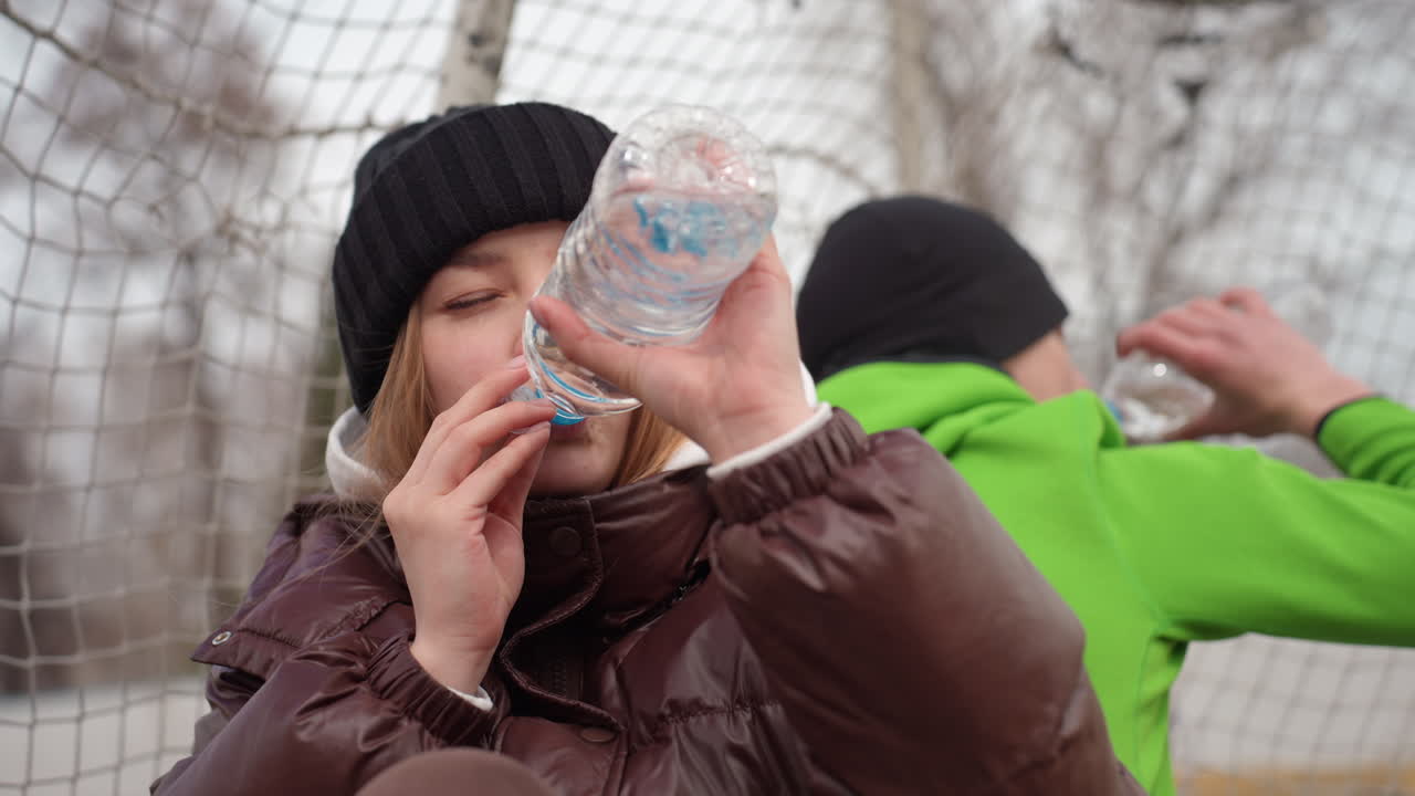 골대 네트 근처에서 검은색 비니를 쓴 백인 여성이 병뚜껑으로 조심스럽게 홀짝이는 모습이에요. 클로즈업으로 집중하는 모습과 차가운 입김을 보여주고, 차분한 겨울 톤으로 반사되는 야외 훈련 중 휴식 시간이에요.