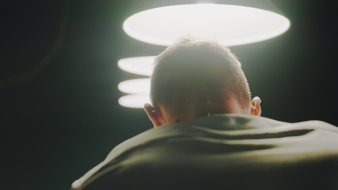 하 셔츠를 입은 남자가 머리 위에 조명과 함께 희미하게 조명 된 방에 서있는 클로즈업 뒷. 그의 실루은 반이는 램프와 대조되어 기분이 좋고 영화적인 분위기를 만니다.