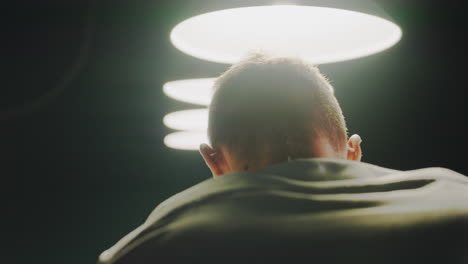 close-up back shot de un hombre con camisa blanca de pie en una habitación débilmente iluminada con iluminación superior. su silueta contrasta con las lámparas brillantes, creando una atmósfera de mal humor y cinematográfica