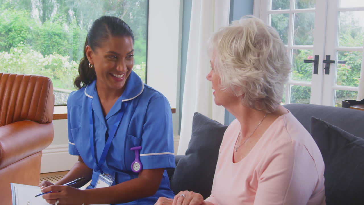 Senior Woman At Home Talking To Female Nurse Or Care Worker In Uniform Making Notes On Clipboard