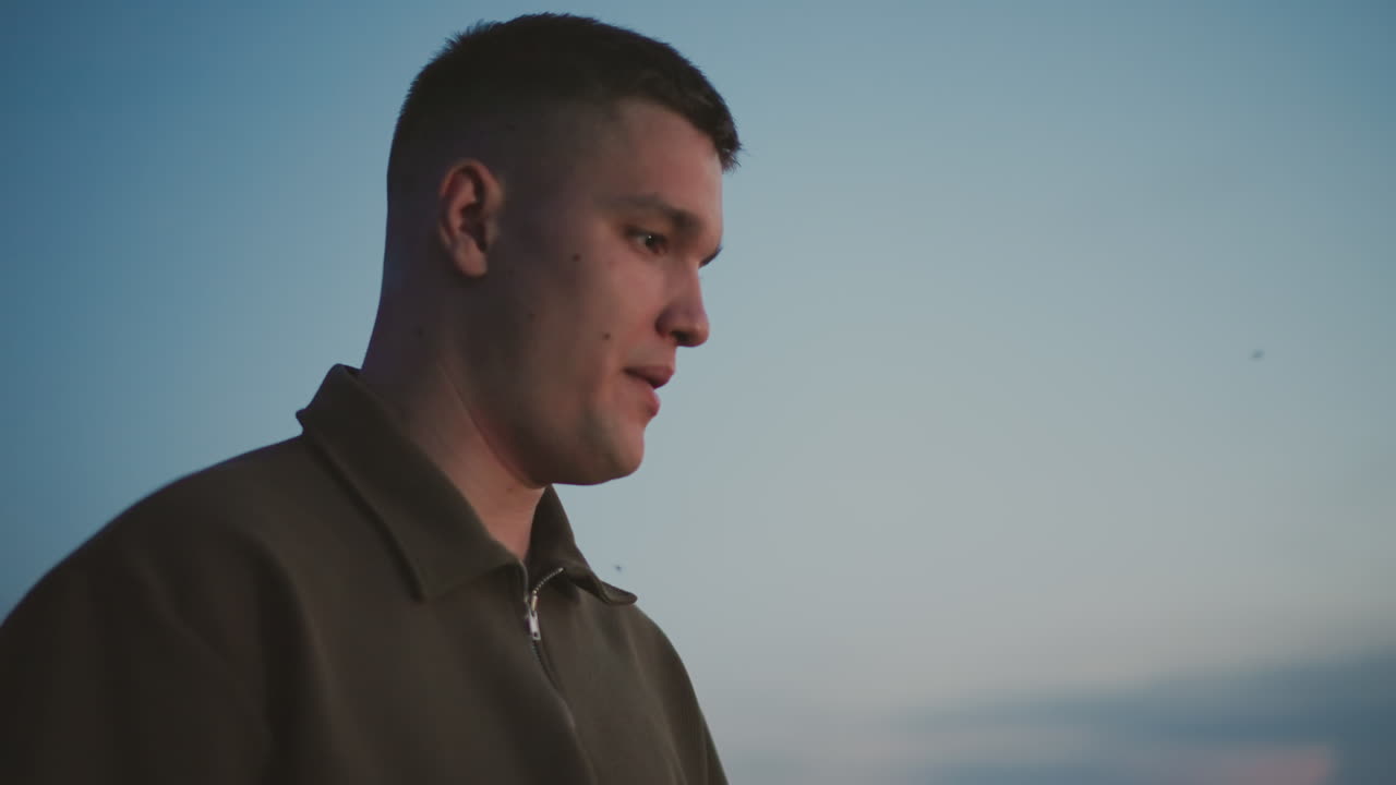 close up of young man standing in grassy field at dusk with insects buzzing around his head under clear sky capturing contemplative expression and tranquil outdoor moment as twilight settles
