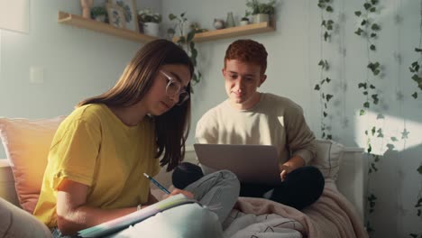 dos adolescentes caucásicos sentados en la cama y aprendiendo de libros y computadoras portátiles juntos.