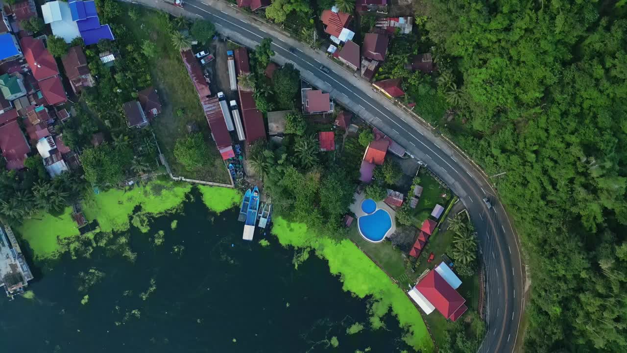 A rotating top-view aerial of the lakeside road winding past colorful rooftops and forested edges in Talisay, Batangas, Philippines