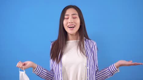 young attractive asian woman stands on a blue background removes a protective medical mask from her face and breathes deeply