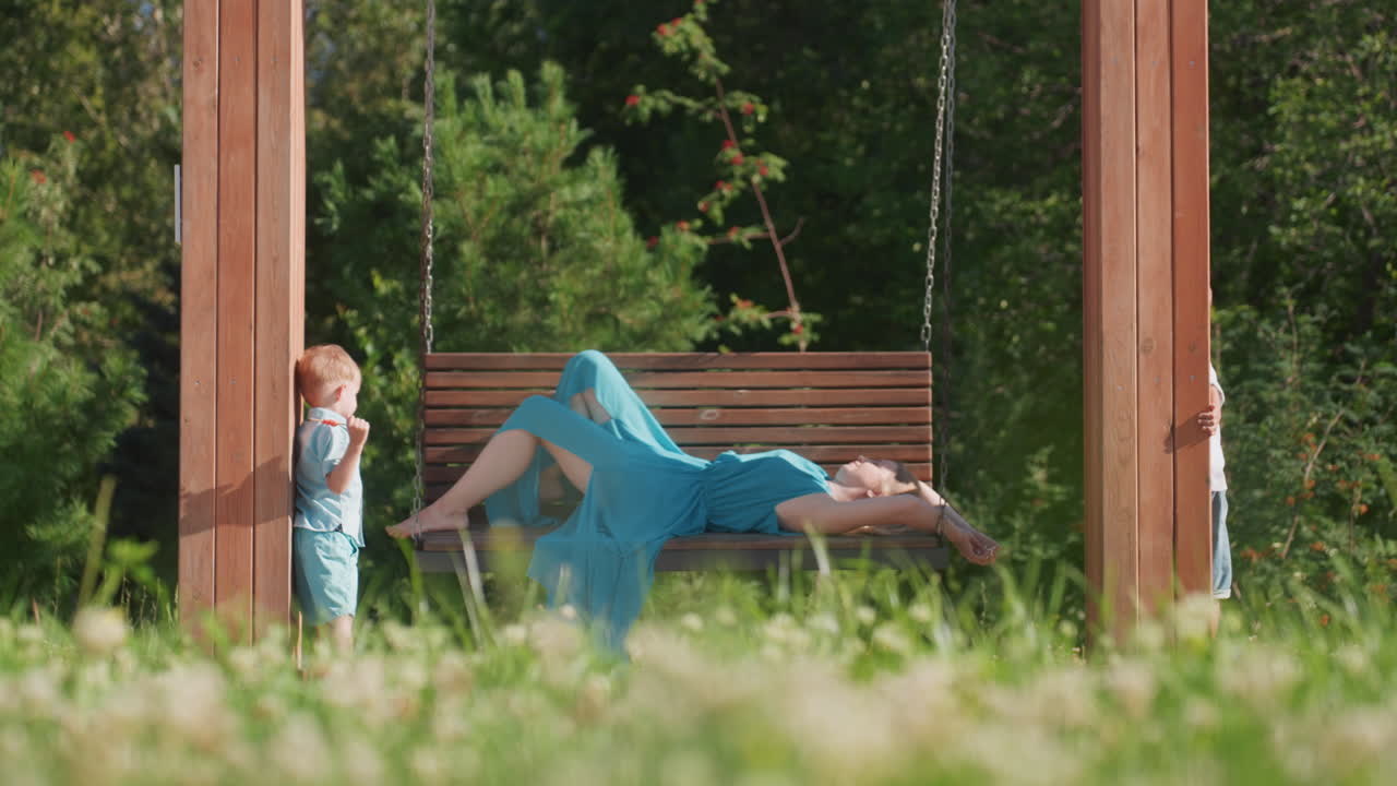 Madre bianca che riposa su un'altalena del parco, il figlio curioso si avvicina, pergola in legno, erba e trifoglio illuminati dal sole, vestito blu svolazzante, momento familiare spontaneo del fine settimana, interazione giocosa, calda atmosfera estiva.
