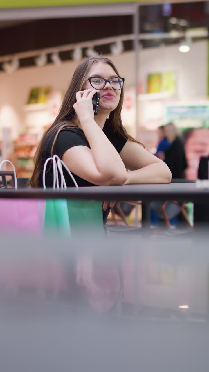 joven elegante con gafas en el teléfono, sonriendo felizmente, mirando a la derecha en el concurrido centro comercial de comidas con fondo borroso de estructuras modernas, mesas, sillas, y un grupo de personas caminando