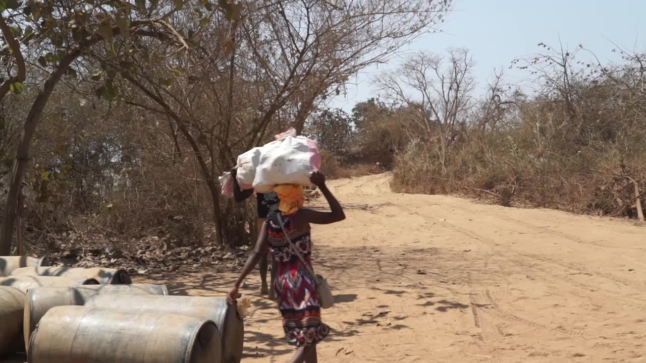African locals balancing sacks on their head to transport along dirt road. Wide shot along road.
