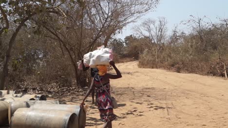 african locals balancing sacks on their head to transport along dirt road. wide shot along road.