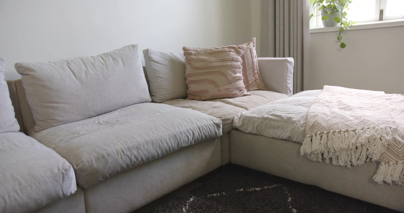 En lysebeige L-formet sofa modtager dagslys fra et gardinbeklædt vindue, hvilket skaber diskrete skyggeforskydninger.