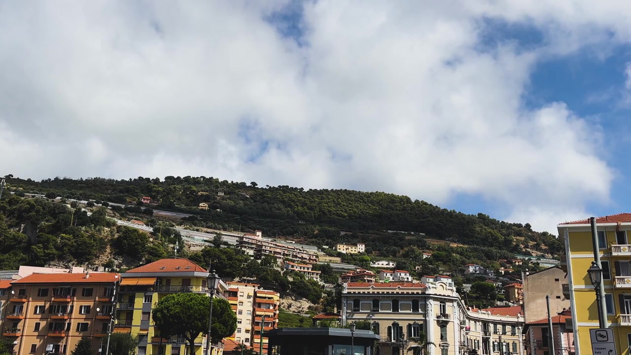 Paesaggio urbano panoramico di Ventimiglia con edifici colorati, tetti in terracotta, colline verdi e nuvole drammatiche che si diradano in un cielo azzurro brillante