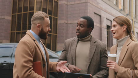 two caucasian and african businessman and caucasian businesswoman talking about their work in the street in autumn while using a tablet