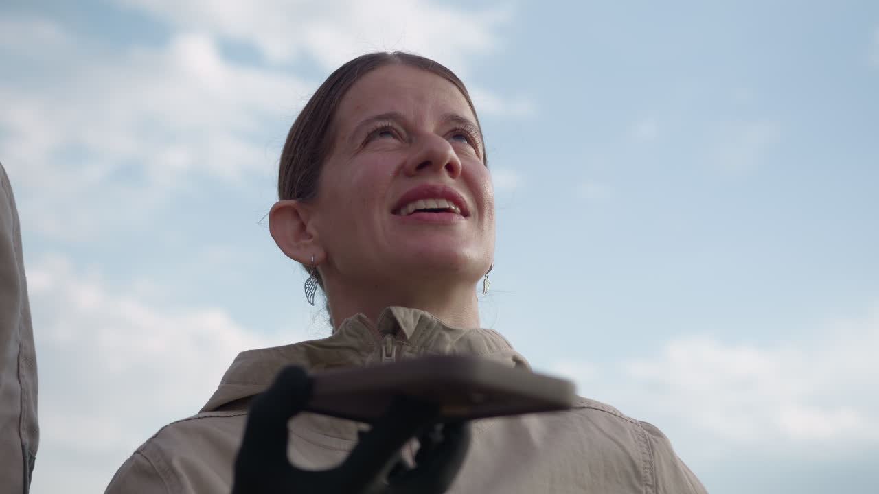 woman gazing into sky while holding device in gloved hand, face illuminated by sunlight, braided hair and dangling earrings set against blue sky and blurred trees, expression focused anticipation