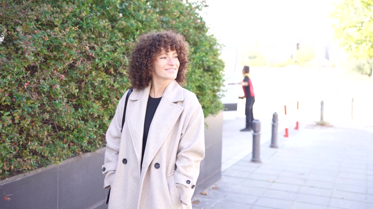 Una donna elegante vestita con un trench in piedi elegantemente tra una vegetazione lussureggiante in un contesto urbano.