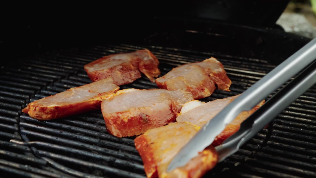 Tykke marinerede kødskiver ligger på mørke grillriste, mens metaltænger justerer stykkerne over varmen