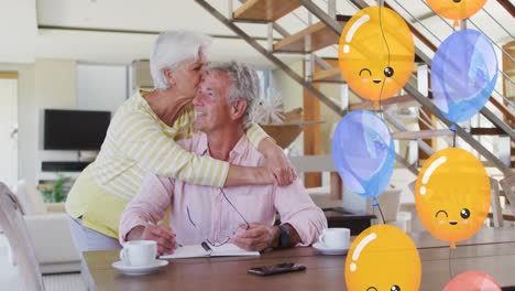 Animation-Von-Partyballons-über-Einem-Glücklichen-Kaukasischen-Seniorenpaar,-Das-Sich-Zu-Hause-Im-Esszimmer-Umarmt