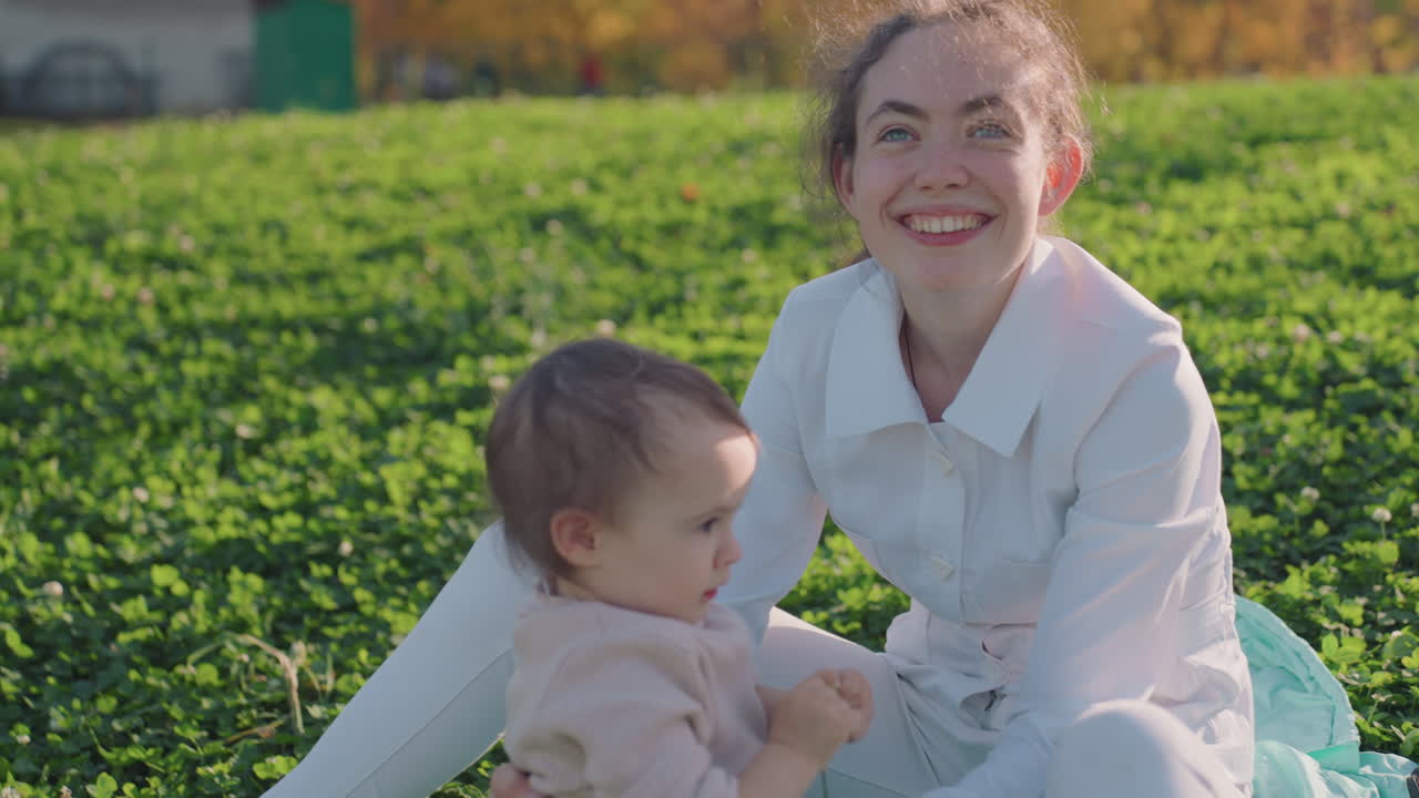 Weiße Mutter und Kleinkind sitzen auf Gras, sonnenbeschienenes Parkpicknick, enge mütterliche Interaktion und sanftes Spiel, weiche Decke und Klee, ein authentischer Moment aus dem Familienalltag, der eine fürsorgliche Bindung und ein warmes Lächeln zeigt.
