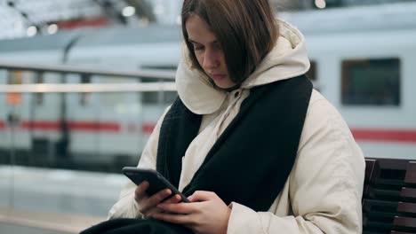 medium shot of casual girl in down jacket with scarf thoughtfully using cellphone waiting at subway station