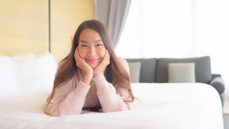 a young, very cute woman lying on a comfortable hotel suite bed has her chin propped up in her hands and smiles