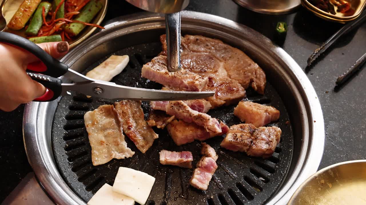 Hand uses scissors to cut pork belly on tabletop grill, bright lighting, overhead camera angle