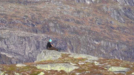 piloto de drones en el pico de la montaña desplegándose y preparando drones de cámara para fotografía de paisajes - hombre caucásico sentado solo en una roca mientras prepara su drone - fondo de montaña borroso noruega