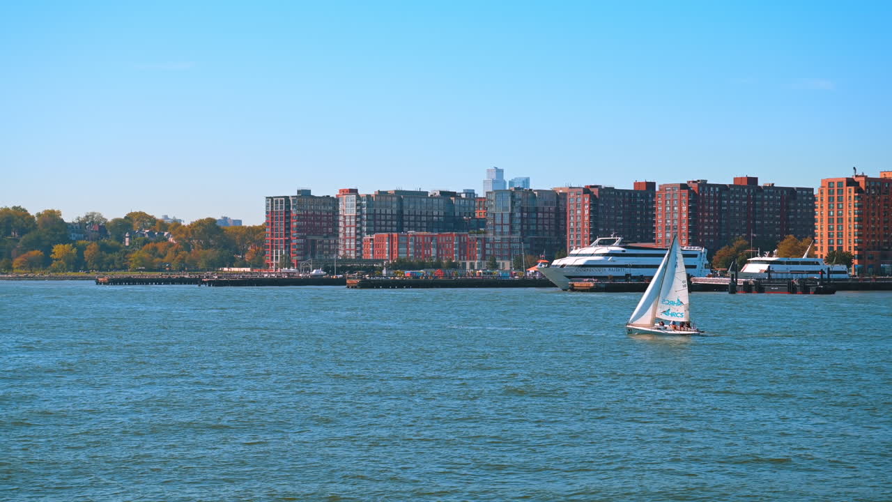New York, USA, 1 novembre 2025: barca a vela bianca che supera un traghetto passeggeri più grande di fronte all'architettura della costa del New Jersey. Ampia veduta delle imbarcazioni sull'Hudson River con gli edifici residenziali in mattoni colorati visibili sullo sfondo sotto un cielo blu