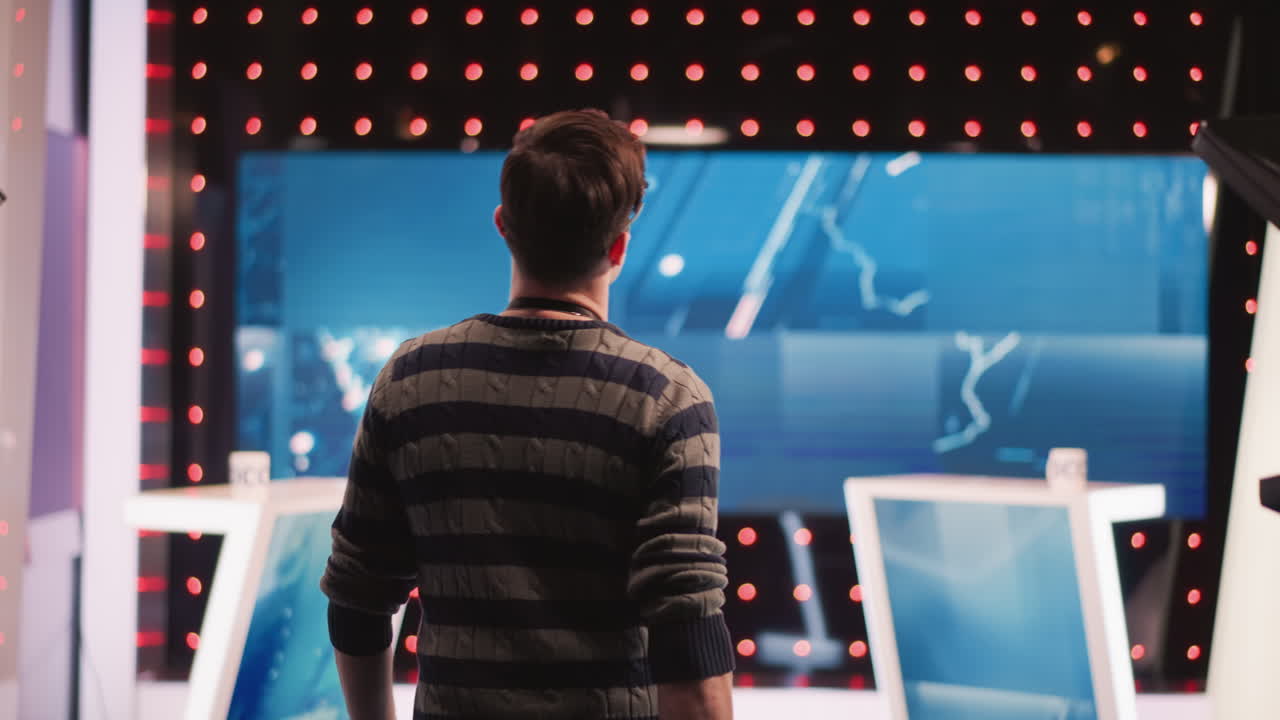 Man adjusting boom microphone in professional studio with neon lights, large cameras, and equipment visible, focusing on sound recording process during video production setup