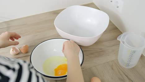 close up slow motion of person splitting a raw egg by hand, dropping yolk and white into bowl, surrounded by empty mixing bowls, on a wooden kitchen surface during cooking session, making tiramisu