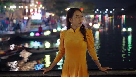 Young-Vietnamese-woman-in-yellow-dress-looking-off-screen-with-night-river-reflections-in-background,-Hoi-An,-Vietnam