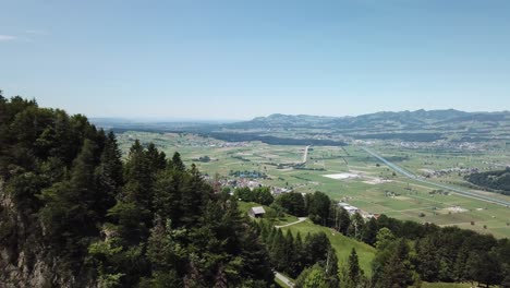 drone flight over a mountain chain or cliff with some trees on it, to show a big valley with some villages and sheds on a small hill in front of the lake of zurich