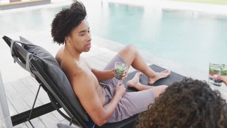 video of happy diverse couple on holiday relaxing with drinks in sun and talking by swimming pool