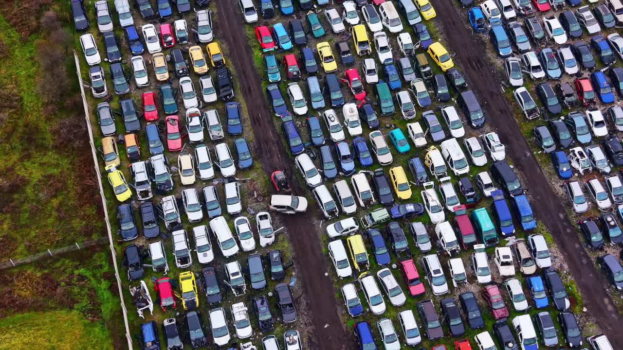 Biler parkeret på en stor parkeringsplads med rækker af køretøjer i forskellige farver og modeller nær et græsareal på en overskyet dag.
