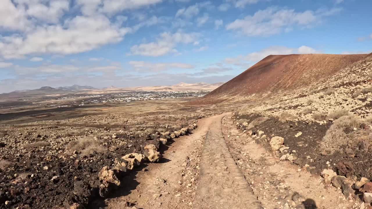 Time lapse of a rugged mountain trail winds through a volcanic landscape, with dark lava rocks and dry vegetation. The path leads towards a distant village under a blue sky with scattered clouds.