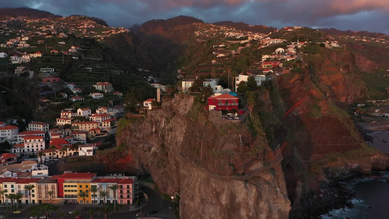 Ripresa con drone che si allontana rapidamente dall'hotel rosso sulla scogliera di Ponta do Sol, sulla costa di Madeira, al tramonto, con drammatiche nuvole temporalesche dietro la montagna.