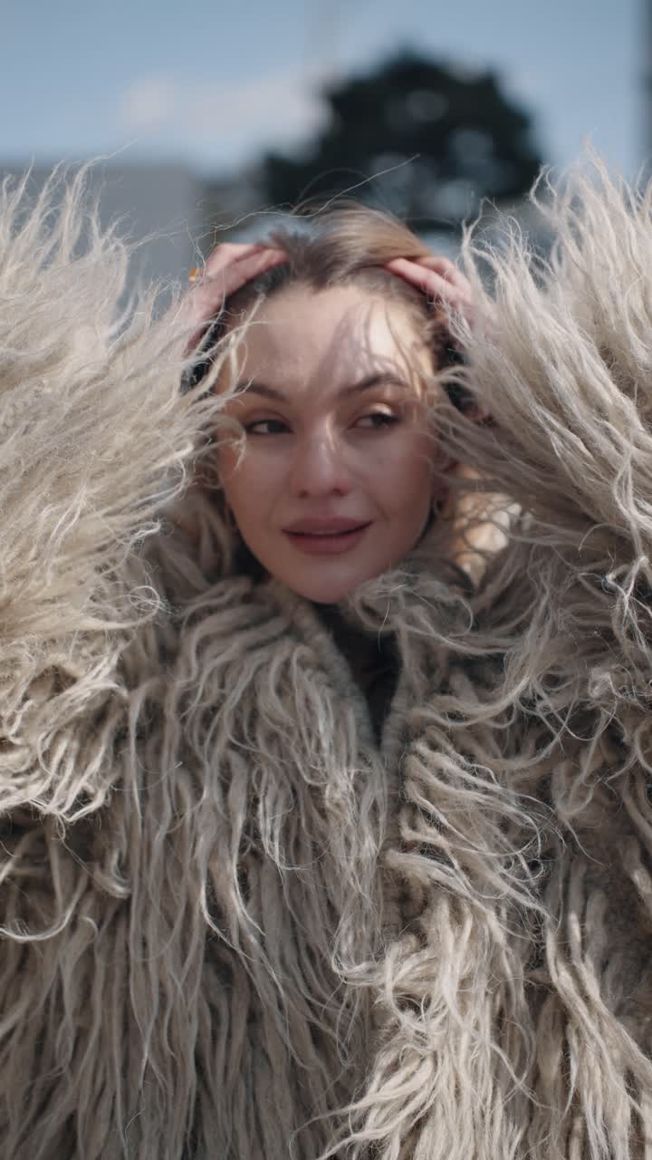 A portrait of smiling fashionable woman brushing hair with hands and wearing a shaggy beige coat stands outdoors in a breezy setting, hair flowing in the wind against a blurred background - vertical