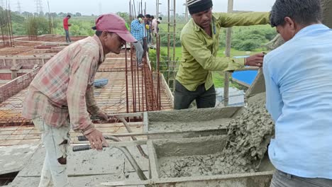 one worker is operating a concrete mixer, while others are guiding the concrete into place. the site has visible rebar structures and scaffolding, indicating that the building is in the early stage