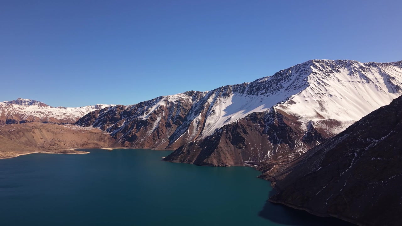 Ampia vista panoramica di un profondo bacino idrico turchese incorniciato da imponenti creste andine innevate e terreno accidentato sotto un cielo invernale limpido e terso. Embalse del Yeso, Cile.