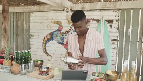 happy african american barman, using tablet and preparing cocktails at beach bar, slow motion