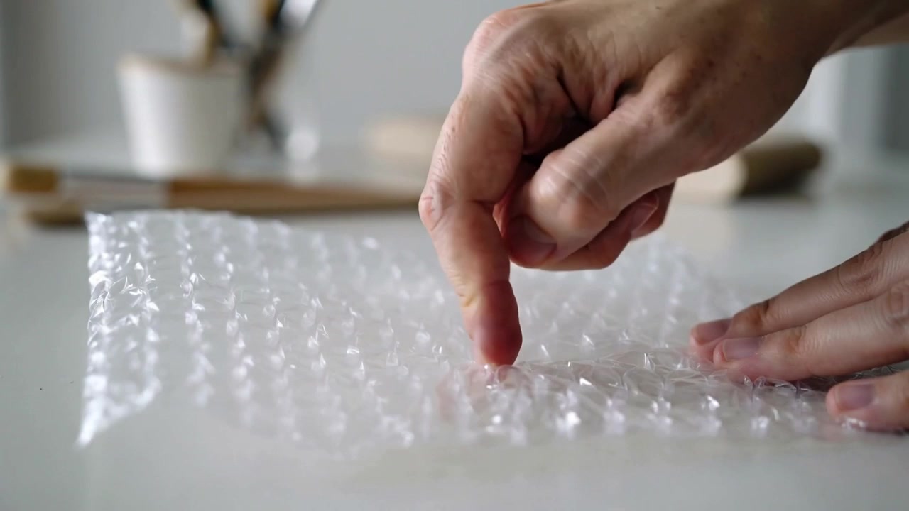 Dito indice che picchietta, mani adulte che premono pluriball trasparente sul piano di lavoro della cucina, facendo scoppiare le bolle.