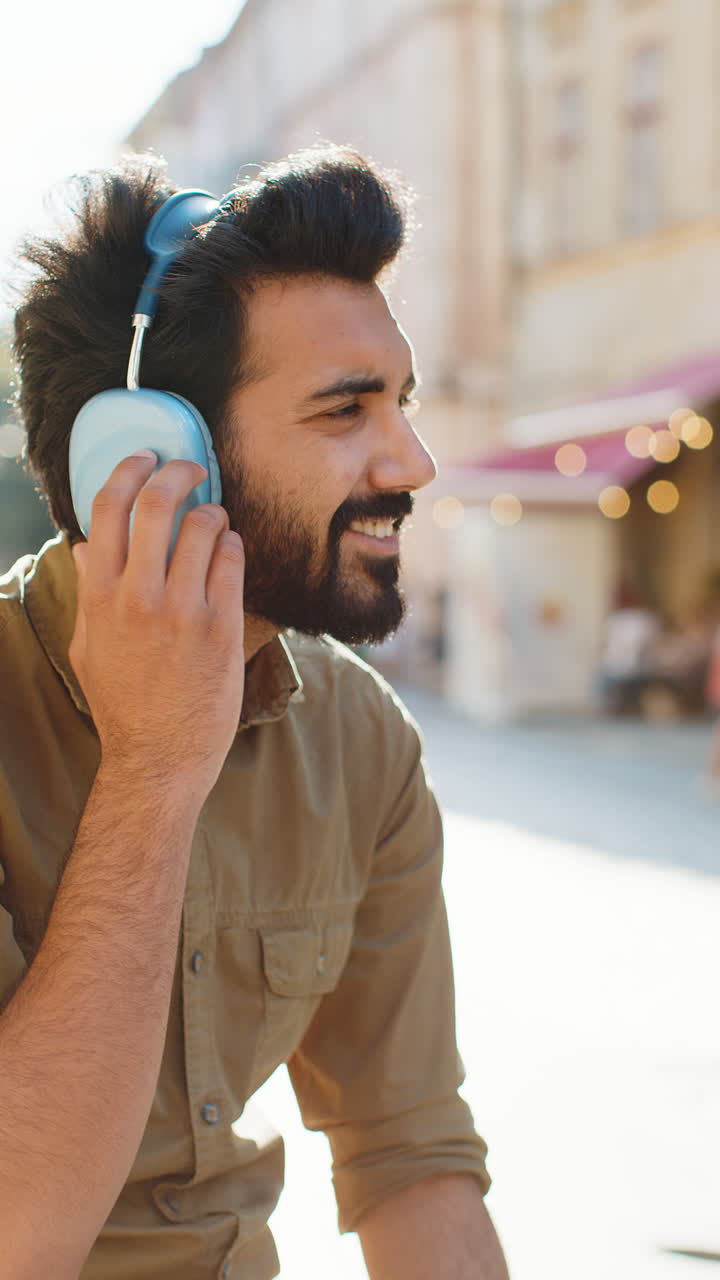 Happy young man in wireless headphones choosing listening music dancing outdoors city street